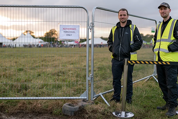 Tall UK security guards stand beside Heras fencing and a retractable barrier marking the perimeter of an outdoor festival, with signage restricting access to authorised personnel only.