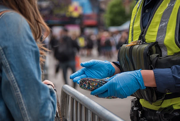 Tall UK security guards checking festival-goers' bags at an outdoor summer event entrance, with a visible sign stating bag search is required for entry.