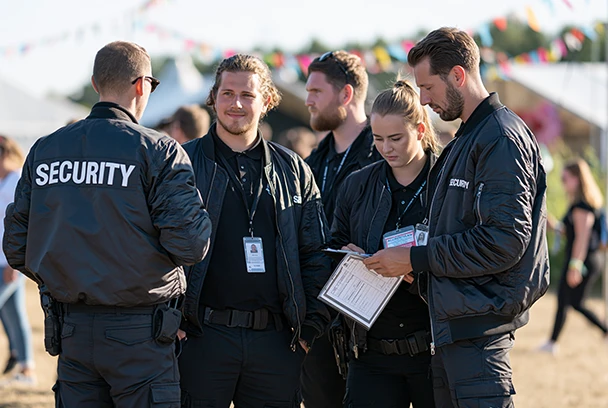 Tall, uniformed UK security guards checking accreditation at an outdoor summer festival entrance