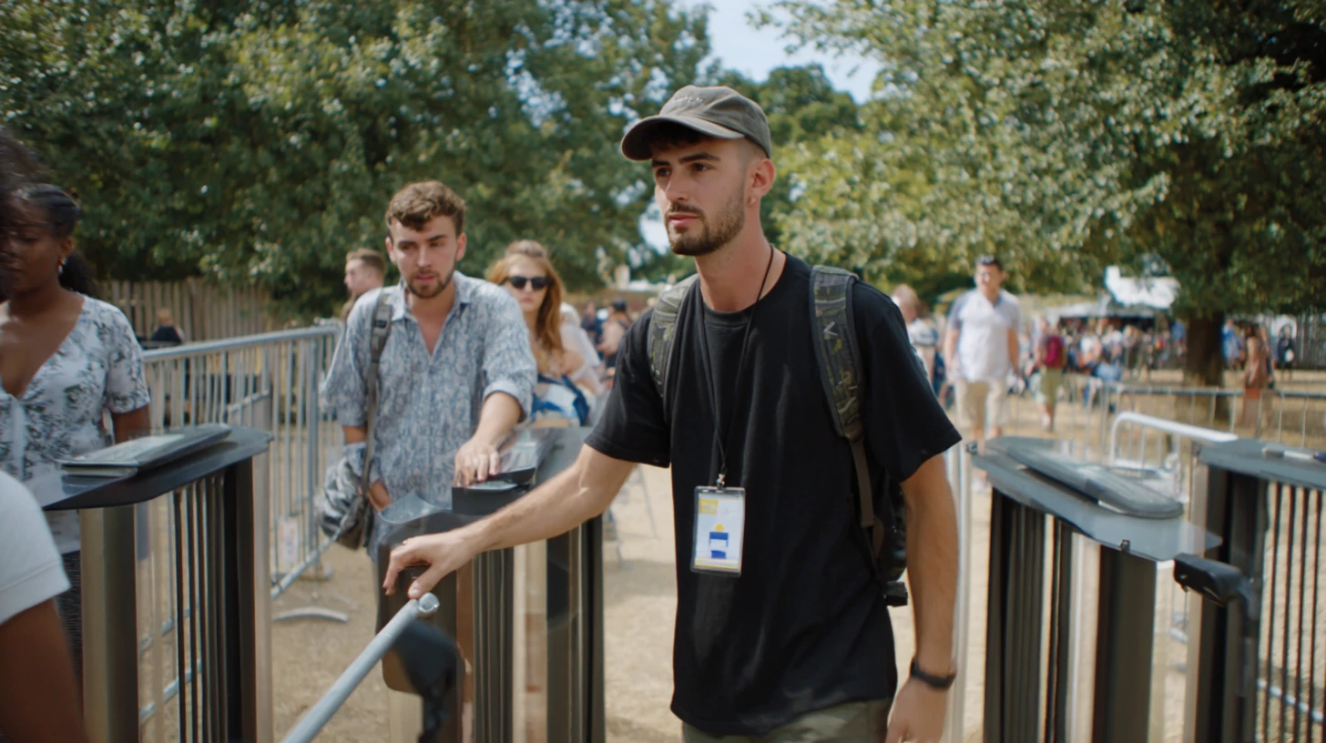 A medium shot of festival goers passing through walk-through metal detector turnstiles at an outdoor event on a sunny day. In the foreground, a young man wearing a khaki cap, black t-shirt, camouflage backpack, and a festival lanyard with a credential badge around his neck pushes through a turnstile gate. Beside him, another young man in a light patterned shirt taps his ticket or wristband on a dark scanning pad mounted to the barrier. To the left, a woman in a floral top is partially visible. The turnstile lanes are flanked by silver crowd control barriers with ticket scanning kiosks attached. In the background, a large crowd of festival attendees moves through the venue grounds, surrounded by tall green trees under a bright blue sky.